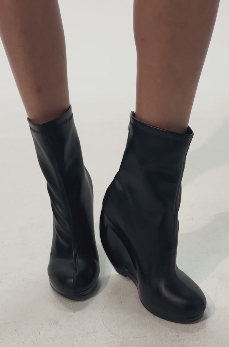 Video of woman modeling the AZALEA WANG RAPTURE OPEN WEDGE COCKTAIL BOOTIE—black faux leather booties featuring sculptural open-cut wedge heels, perfect for making a bold, stylish statement against any backdrop.