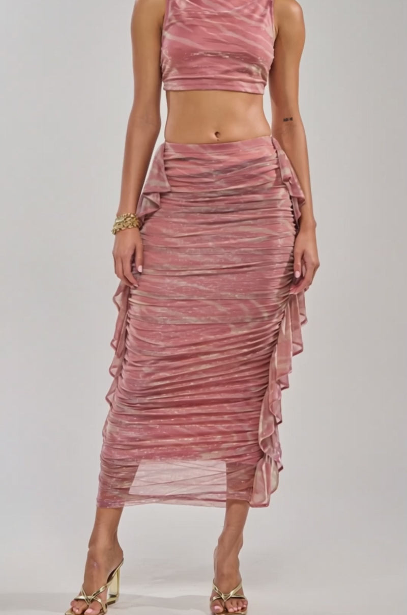 Video of woman modeling the PLAY WITH ME MAXI SKIRT—a high-waisted pink metallic skirt with ruffle and ruched sides. She pairs it with a sleeveless crop top, curly hair, gold hoop earrings, bracelets, and gold heeled sandals for a chic vacation look.