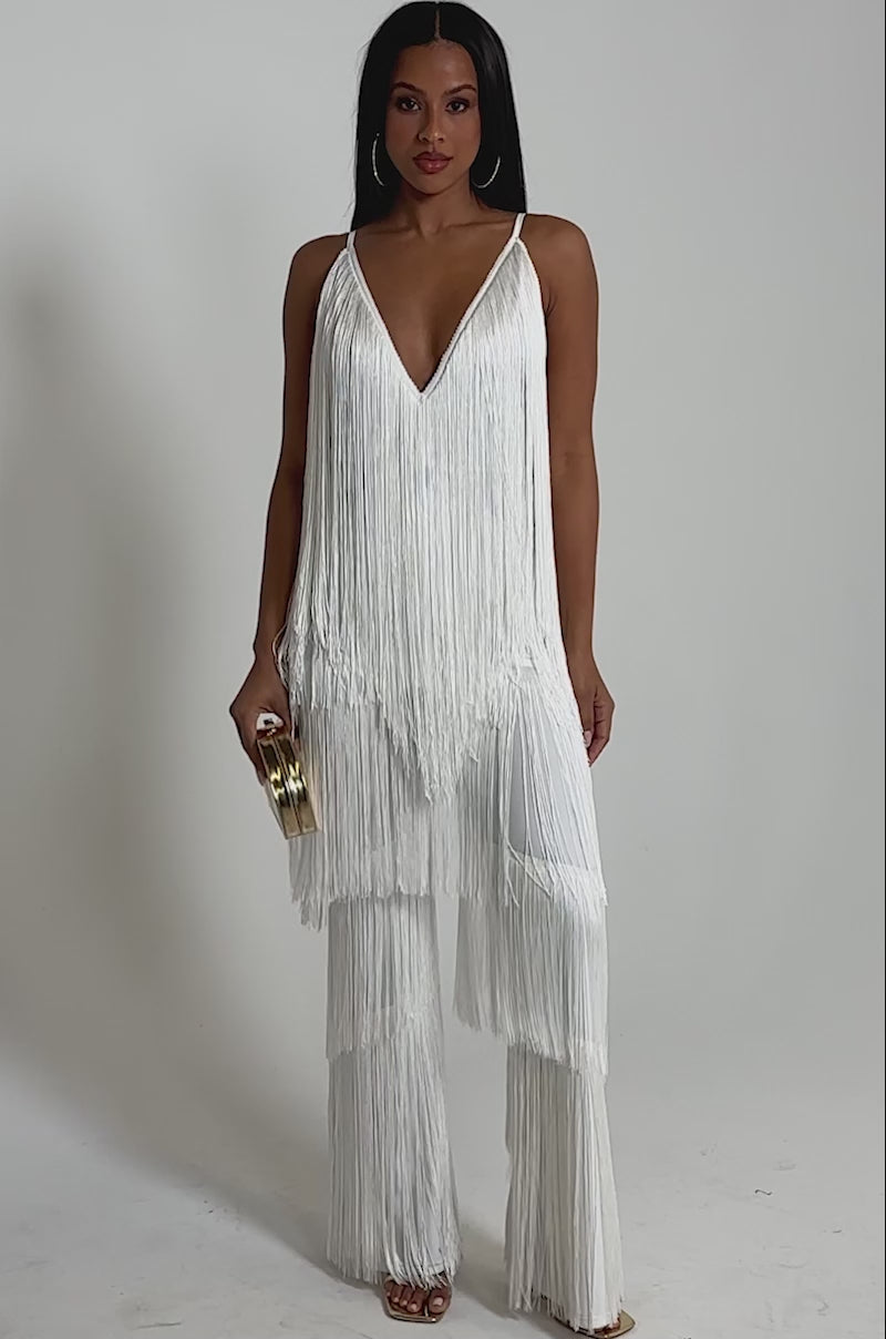 A video of a woman wearing the BAECATION FRINGE JUMPSUIT featuring a sleeveless design, white fringe details, deep V-neckline, and wide-leg pants. 