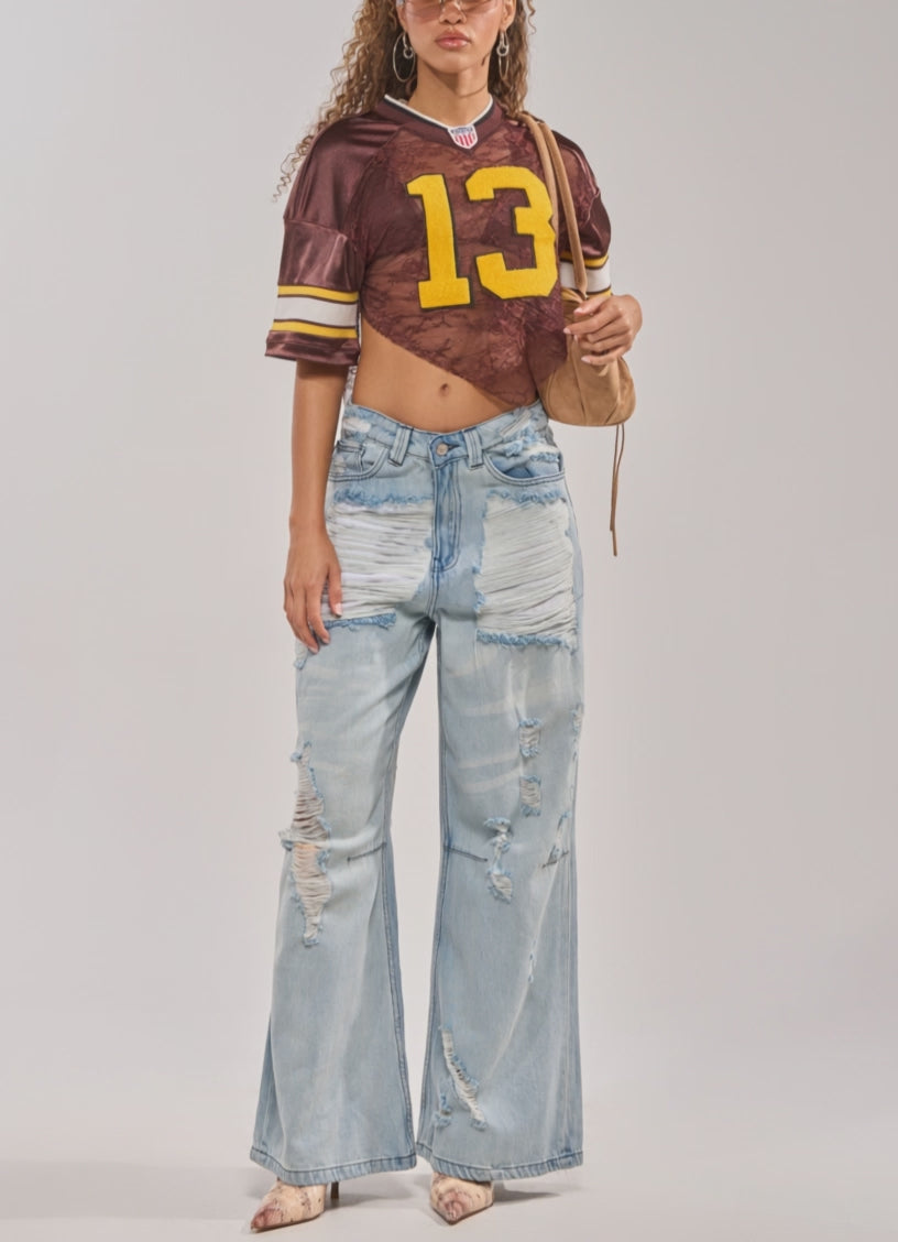 Video of woman modeling the LUCKY LACE JERSEY TOP IN BROWN with varsity patch number 13, sheer lace fabric, paired with distressed light-wash jeans. She accessorizes with hoop earrings, sunglasses, curly hair, and a brown tote bag.