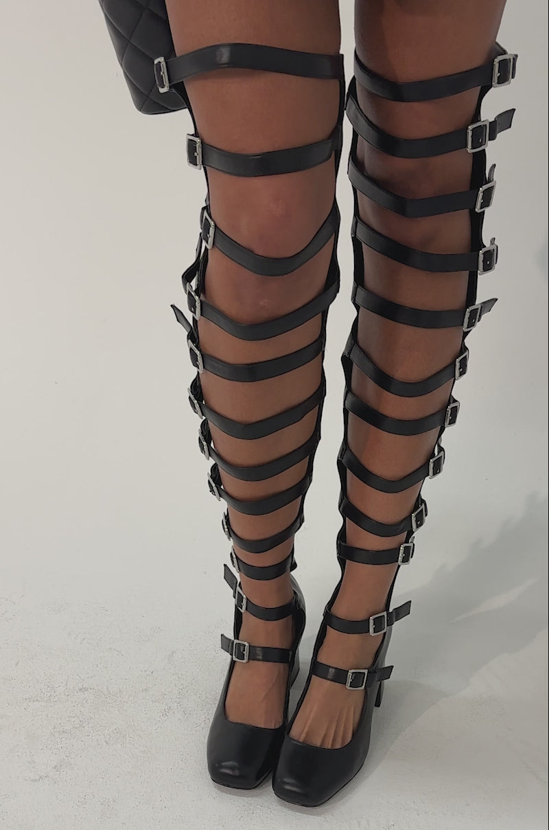Video of woman modeling the AKIRA GOLD YVETTE BLACK OVER THE KNEE MARY JANE BOOT. The black, strappy faux leather boots feature multiple buckles, open sections, a block heel, and reach over the knee.
