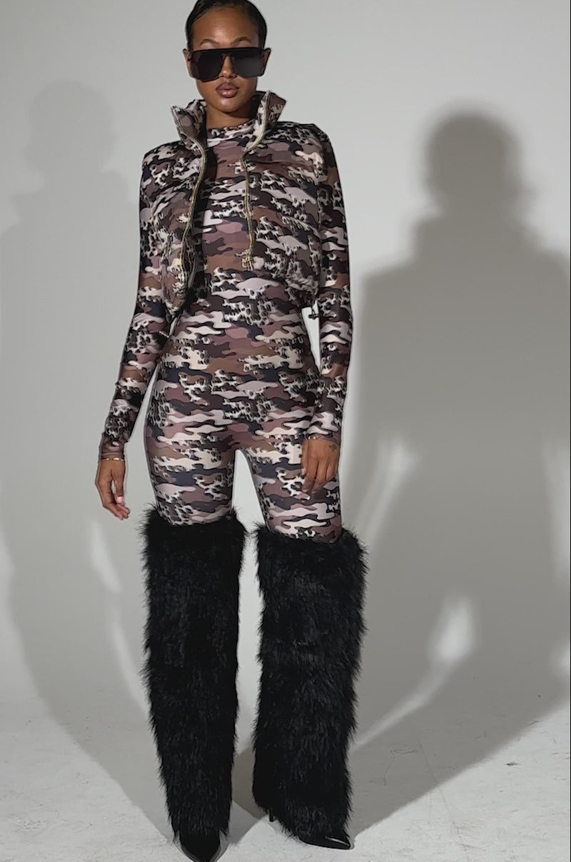 A video of a woman modeling the CAMO CHEETAH PRINT PLAYSUIT, styled with a cropped puffer vest, oversized sunglasses, and black furry knee-high boots against a plain light background.