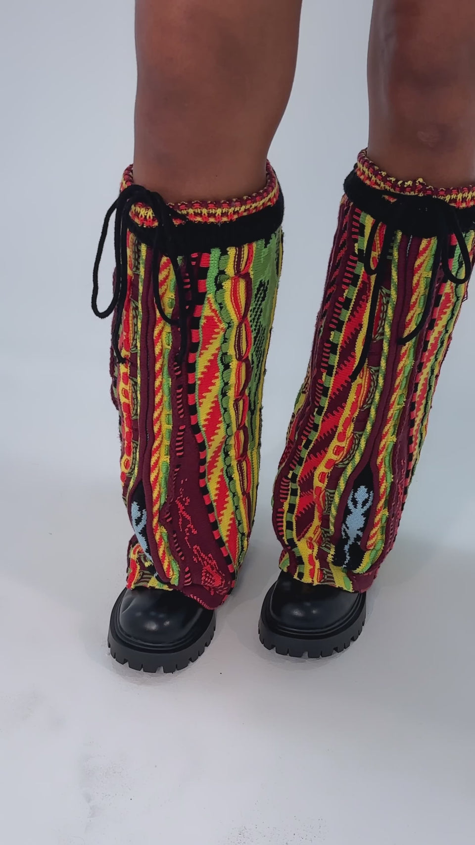 A video of a person wearing the AKIRA GOLD NEIL SWEATER COMBAT BOOTIE—chunky heel faux leather boots styled with colorful, knit leg warmers featuring red, yellow, green, and black patterns—standing on a light background.