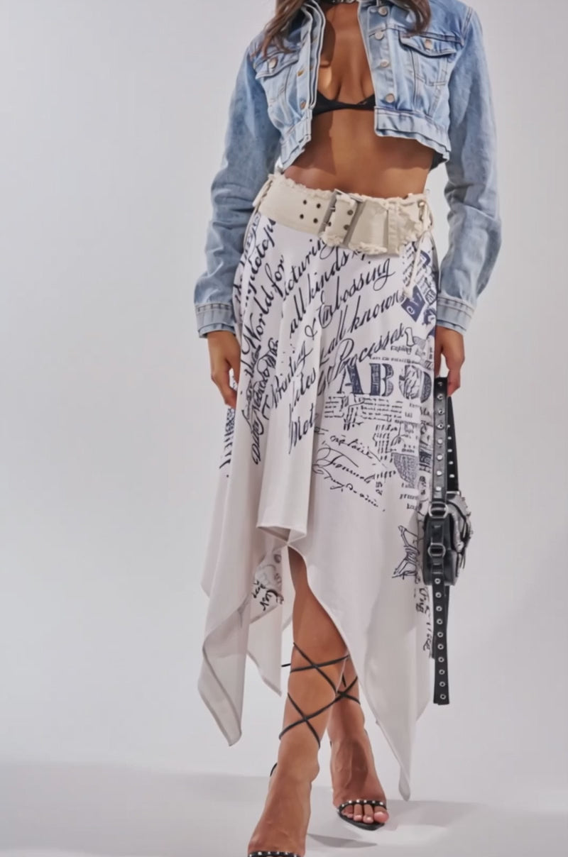 A video of a woman in the NO REPEATS LAYERED DENIM JACKET over a black bralette, pairing it with an asymmetrical white skirt featuring navy script, strappy heels, sunglasses, and a black handbag.
