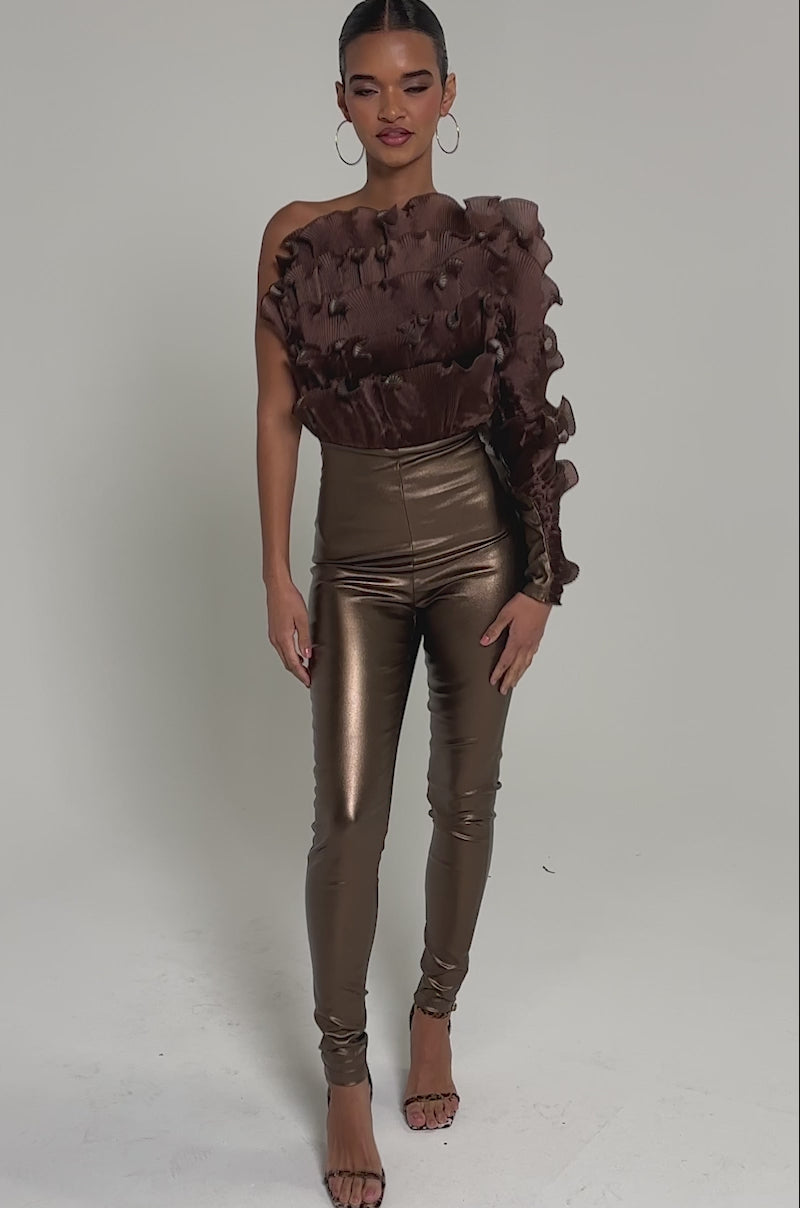 Video of woman modeling the COCOA BUTTER ONE SLEEVE JUMPSUIT, a textured brown metallic piece with one sleeve, paired with heels. Her hair is styled in a long, sleek braid against a plain light background.