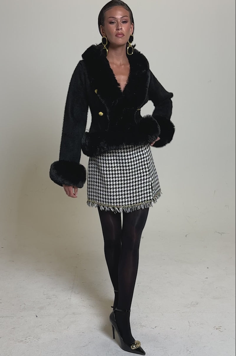A video of a woman posing in the SOFT TOUCH CARDIGAN IN BLACK styled with a houndstooth mini skirt, black tights, gold-tipped heels, large hoop earrings, and slicked-back hair against a light gray background.