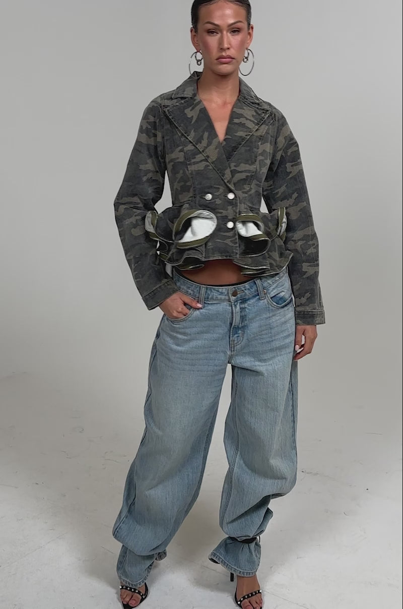 Video of woman modeling the PEPLUM CAMO JACKET, featuring ruffled details, paired with light blue jeans as she poses 