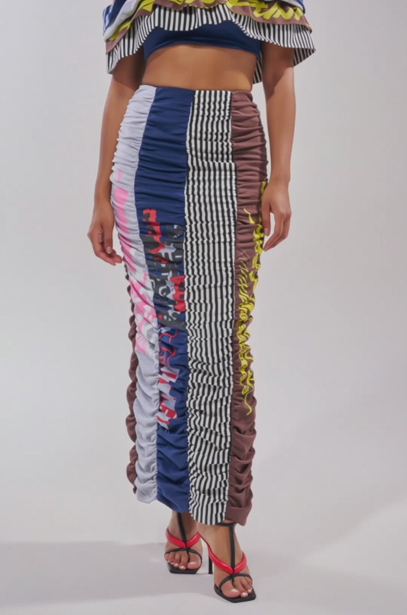 A video of a model wearing the THAT GIRL GRAPHIC TEE MAXI SKIRT, a long patchwork skirt with blue, grey, brown, and black-and-white check panels plus graphic prints, styled with black and red strappy heels.