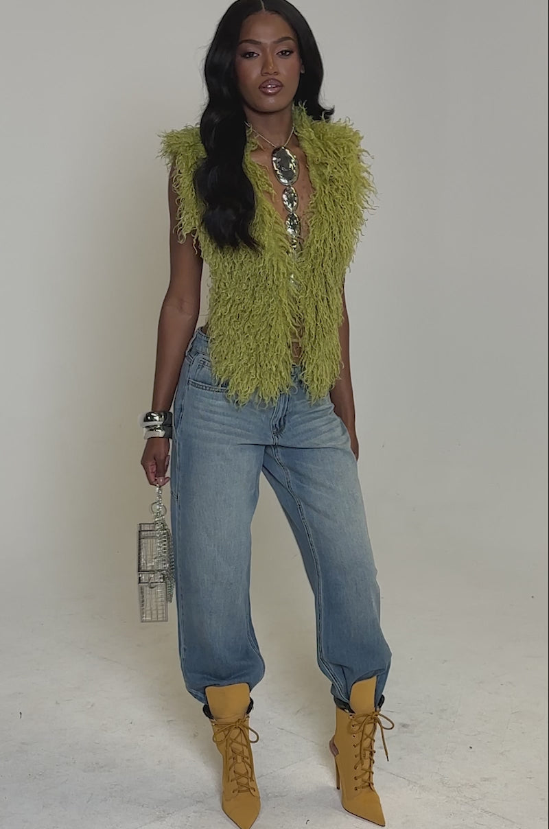 A video of a woman standing against a plain gray background wearing the JAMEELA TOP IN GREEN, light blue baggy jeans tucked into mustard yellow boots, chunky jewelry with silver bangles, and holding a small silver handbag.