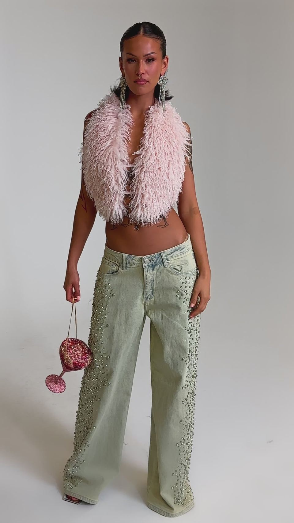 A video of a woman in the FEELING EXTREME RHINESTONE DENIM wide-leg jeans, a fluffy pink vest, and hoops, holding a sparkly pink wine-glass-shaped bag against a plain gray background.