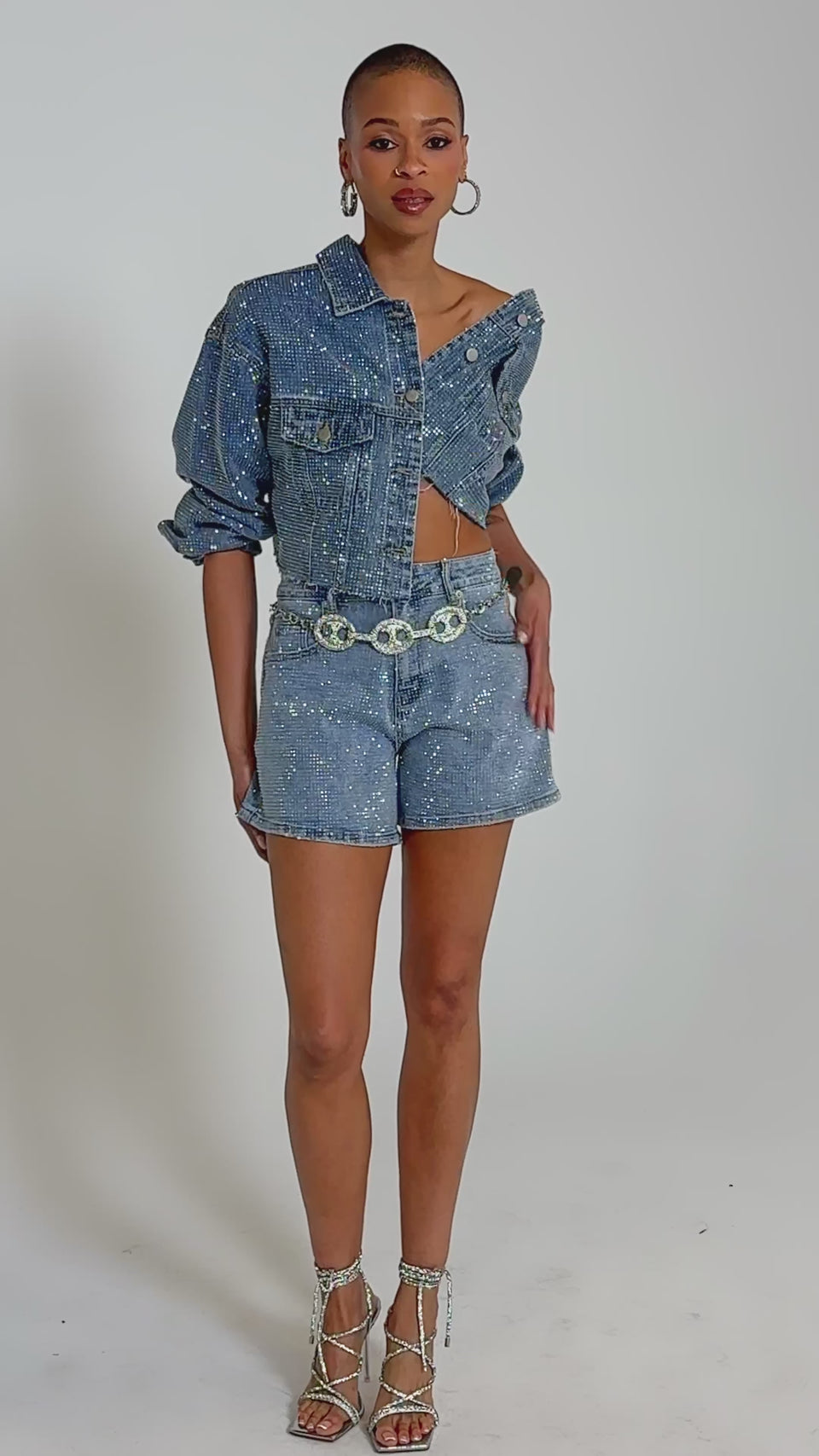 Video of woman modeling the KYLIE EMBELLISHED DENIM JACKET and matching high-waisted shorts, styled with hoop earrings and a silver chain belt featuring circular details