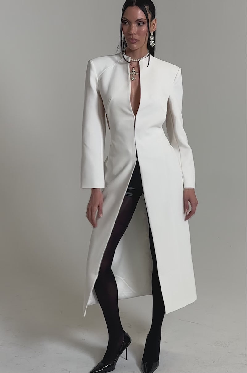 A video of a woman wearing the WHEREVER YOU ARE CUTOUT TRENCH IN WHITE styled with black tights, pointed-toe heels, and a statement necklace.
