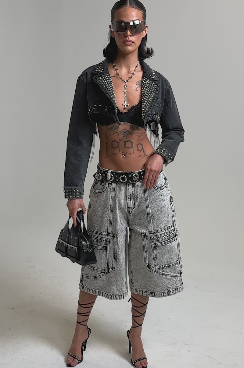 A video of a model wearing sunglasses, a studded cropped denim jacket, black lace bralette, strappy sandals, a small black handbag, and the TROUBLE MAKER BERMUDA SHORT with high-rise waist and cargo pockets.