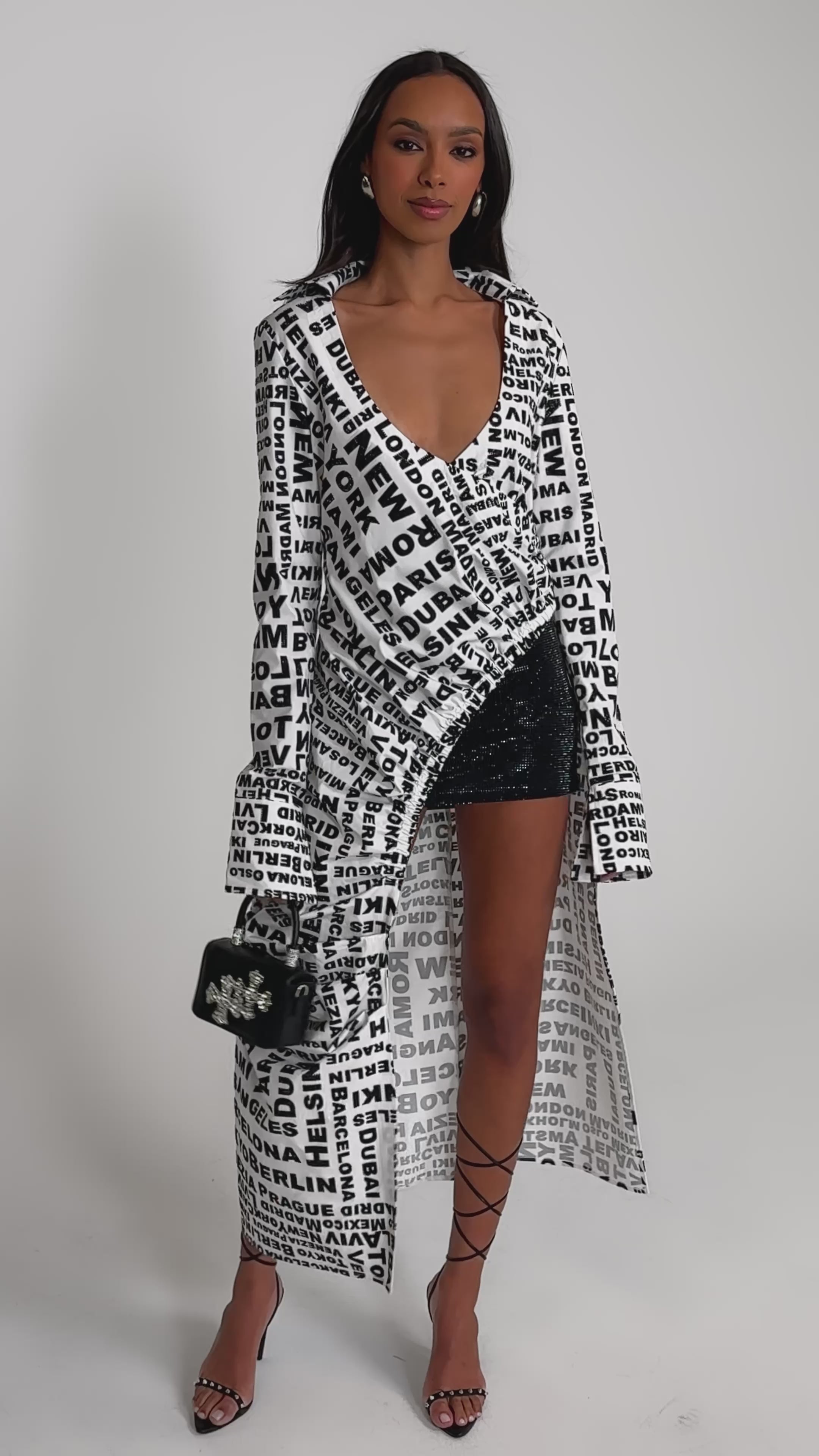 Video of woman modeling the ALL AROUND THE WORLD BLOUSE—a long, white top with typography print—over a sparkly black dress. She accessorizes with a small black purse, strappy high-heeled sandals, and has straight, loose hair.