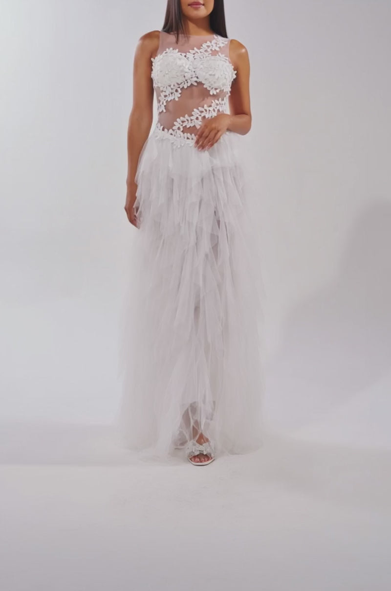 A video of a woman wearing the FELT CUTE RUFFLE MAXI DRESS WITH FLORAL APPLIQUES— a sleeveless, sheer mesh white dress with ruffles and a layered tulle skirt.