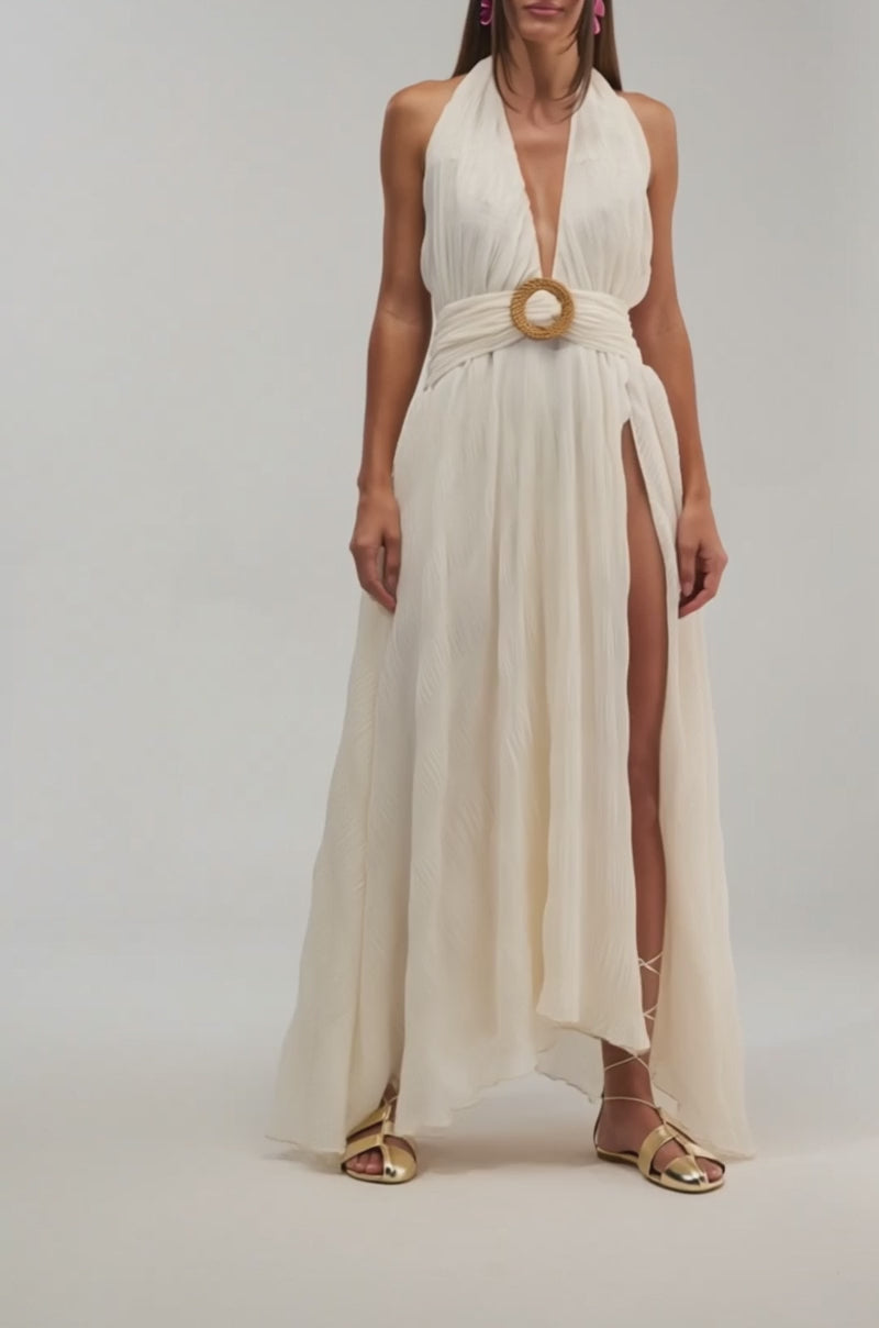 Video of women modeling the BEACH CLUB BELTED MAXI DRESS IN WHITE—a sleeveless halter maxi with pleated chiffon, deep slit, and bold circular belt buckle—with pink earrings, gold strappy sandals, and loose flowing hair for a chic summer look.