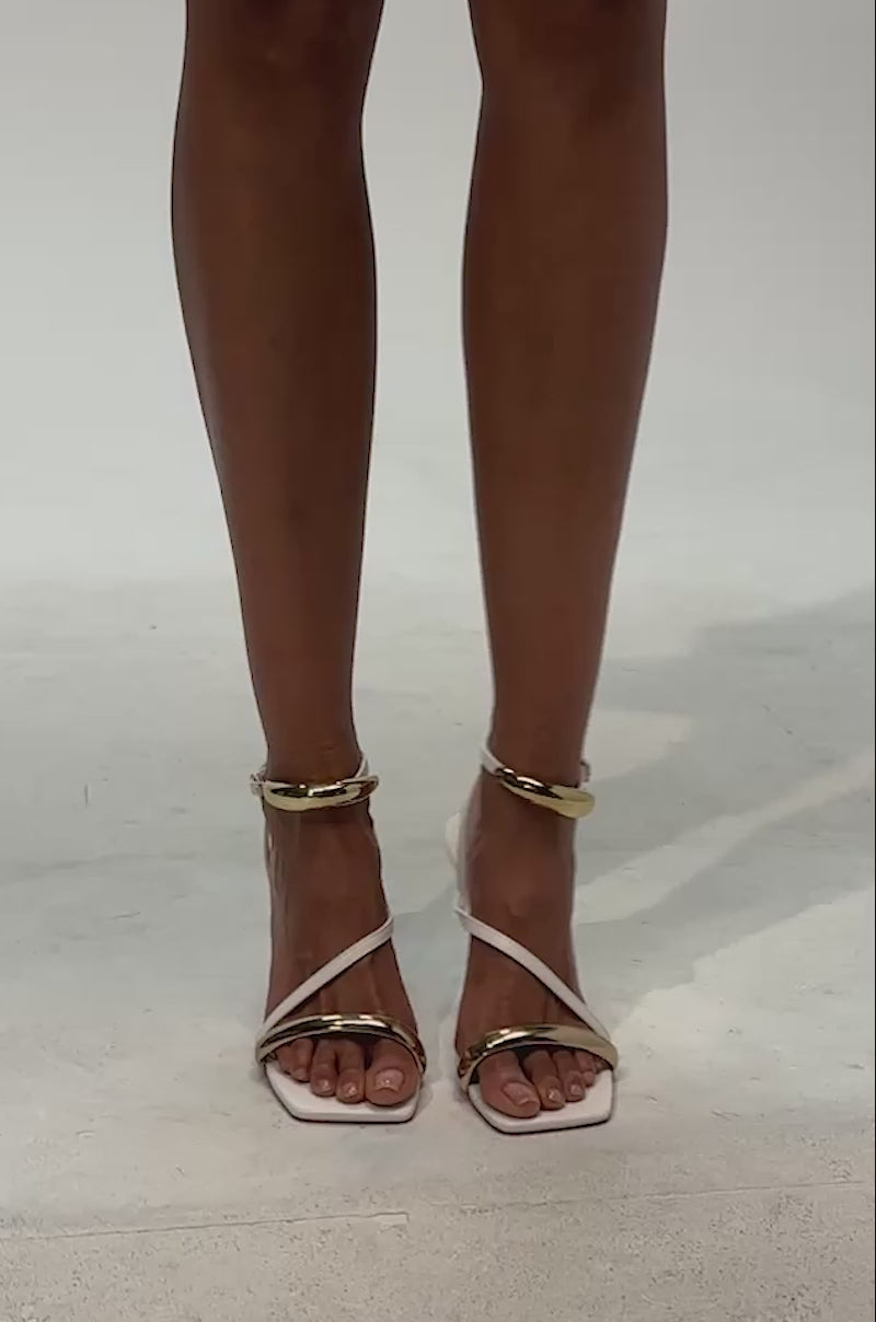 Video of woman modeling the AZALEA WANG WHITE STRAPPY STILETTO AND GOLD HARDWARE SANDAL, featuring gold accents around the ankles and toes, standing on a light background.