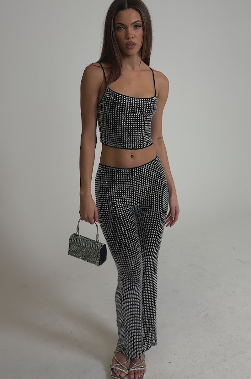 Video of woman modeling the ALL EYES ON ME RHINESTONE FLARE TROUSER set, featuring a matching black crop top and rhinestone flare pants. She accessorizes with a small black handbag and strappy silver heels against a light background.
