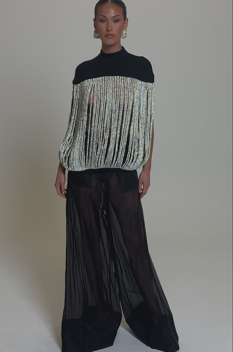 Video of woman modeling the DRIPPING IN DIAMONDS BLOUSE—a black top featuring long silver rhinestone fringe that reveals her torso—styled with sheer black pants and bold earrings, her slicked-back hair set against a plain gray backdrop.