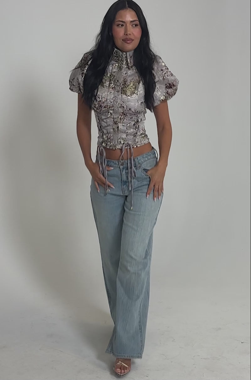 Video of woman modeling the DARLING FLORAL BROCADE PUFF SLEEVE BLOUSE, which has a corset-inspired bodice and lace-up front, paired with light blue jeans against a plain light background.