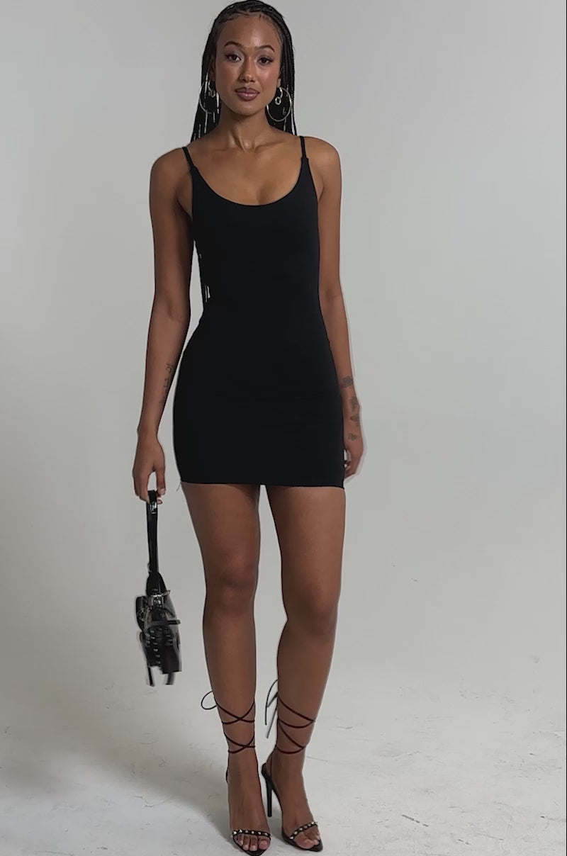 A video of a woman in the REYNA MINI BODYCON DRESS IN BLACK, lace-up heels, hoop earrings, and holding a small black handbag.