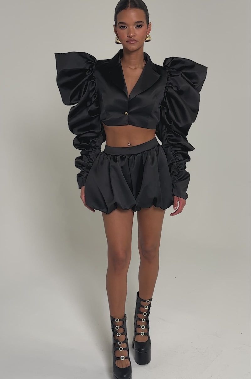 Video of woman modeling the PHOEBE SATIN BUBBLE SHORT. She completes the look with platform boots featuring circular cutouts and large hoop earrings.