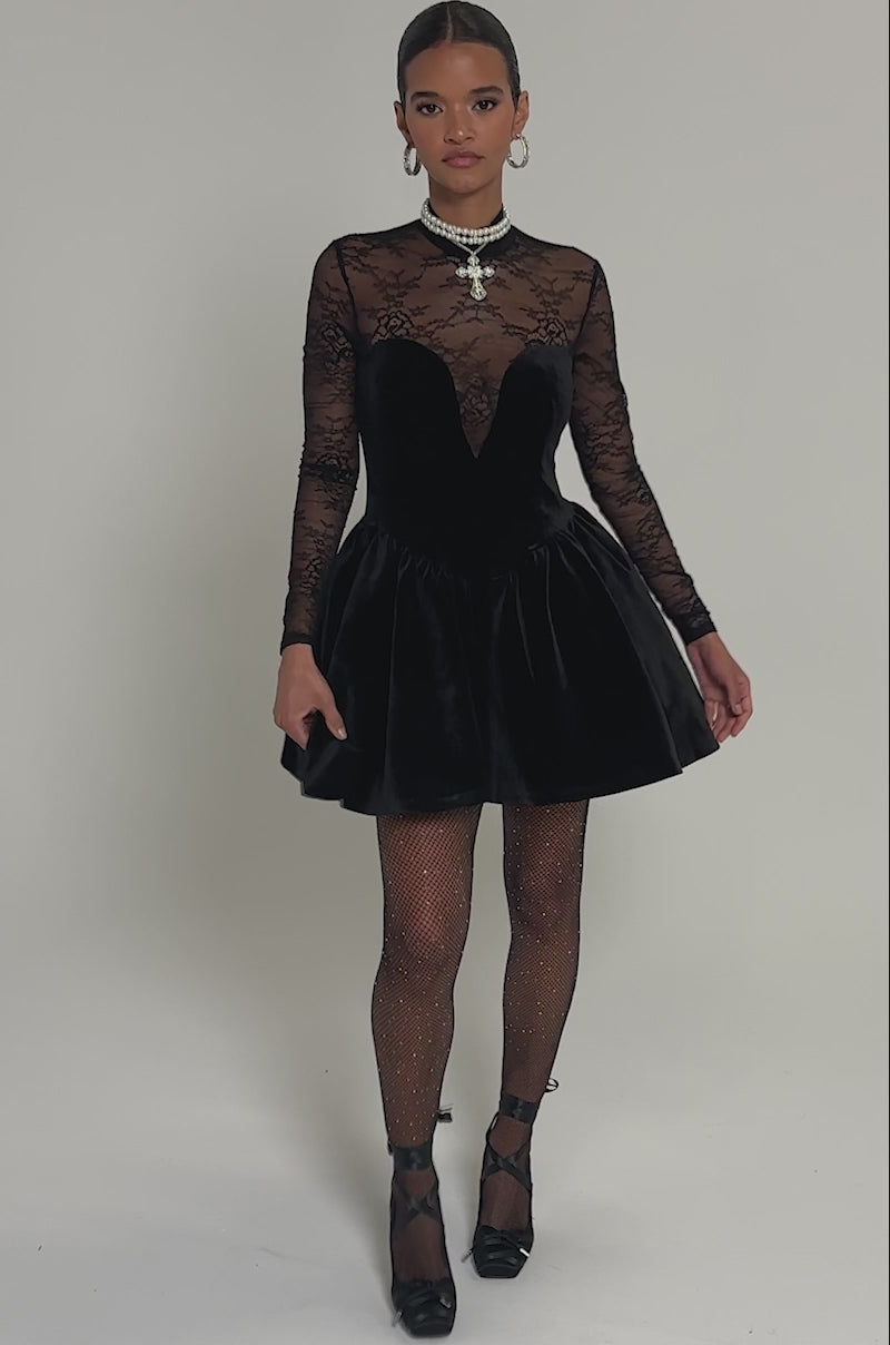 Video of woman modeling the DRESSED TO IMPRESS LACE MINI DRESS, featuring a velvet LBD with a floral lace long-sleeve bodice, styled with rhinestone fishnet tights, black heels, and layered necklaces against a plain background.