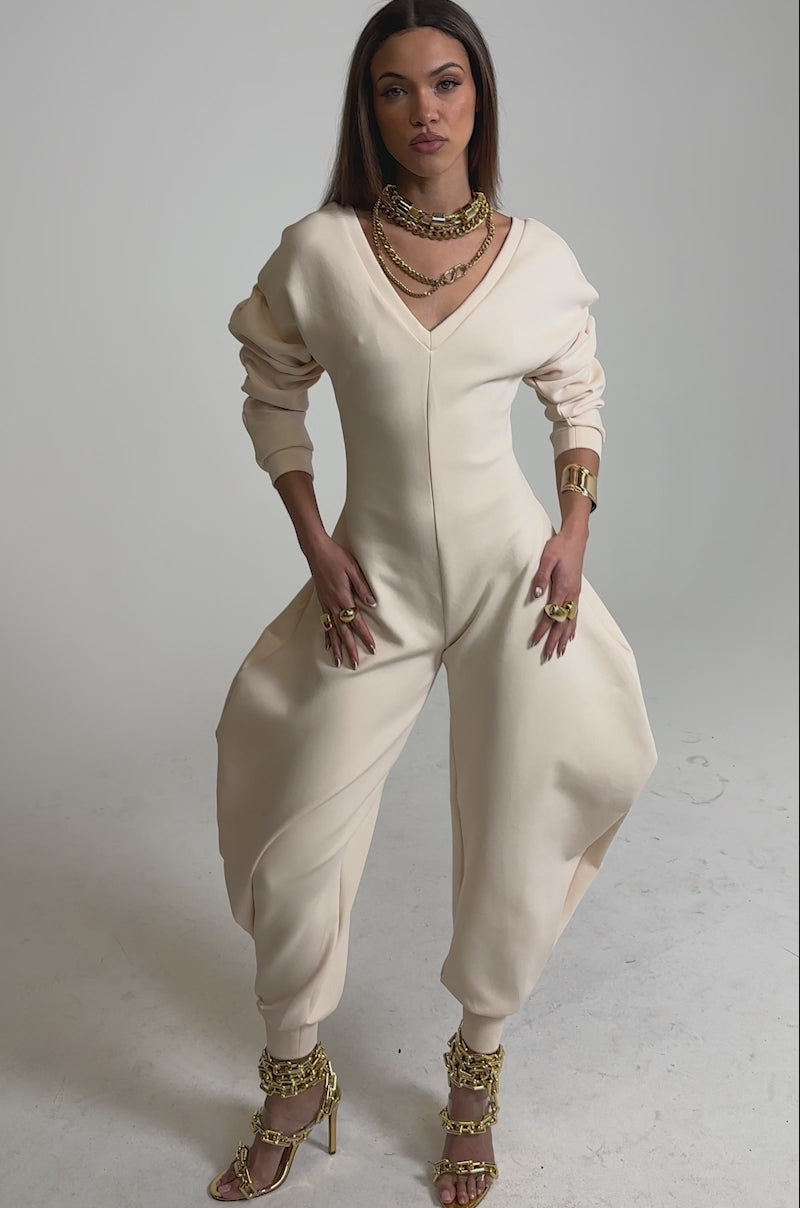 Video of woman modeling the COMFY COZY SWEATSHIRT JUMPSUIT IN BONE, featuring a cream hue and relaxed fit. She accessorizes with chunky gold jewelry and high-heeled sandals, blending comfort with chic style against a plain background.