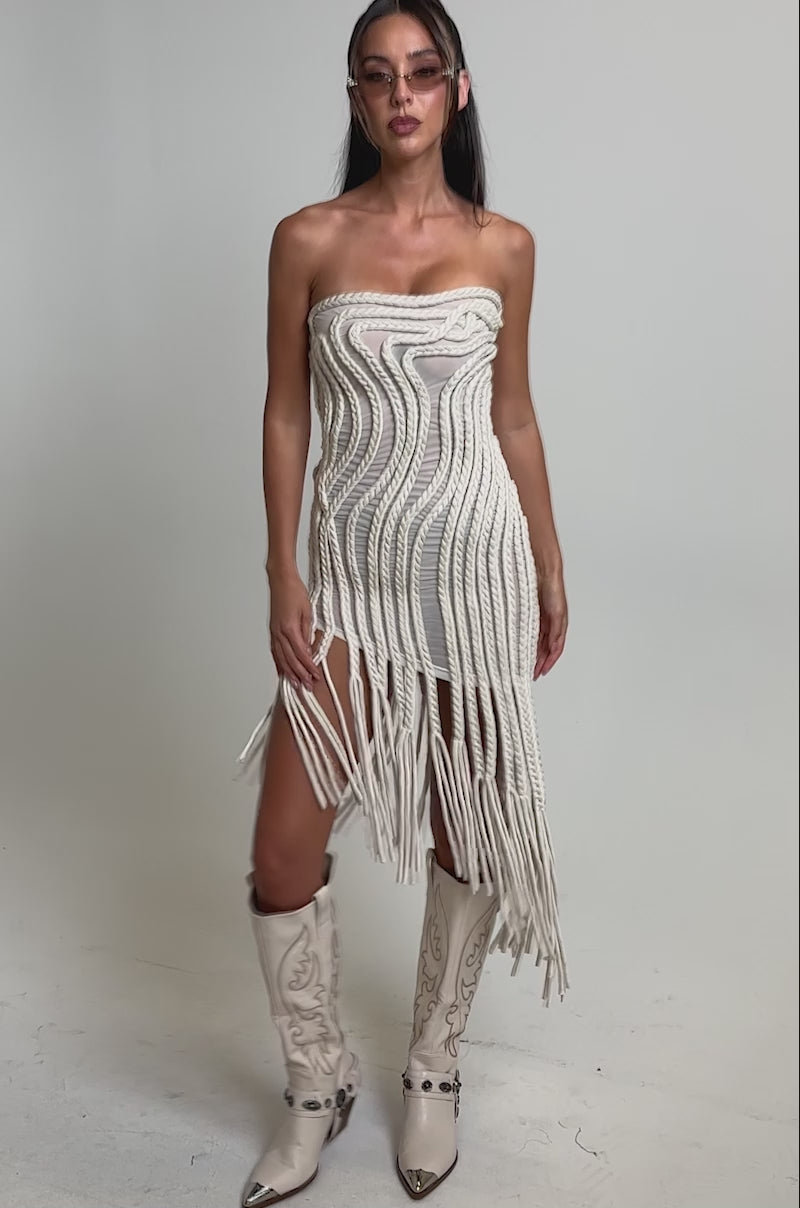 A video of a woman wearing the ALL OVER THE PLACE BRAIDED MIDI DRESS IN IVORY, featuring a sculptural wave pattern and long fringe, paired with white cowboy boots.