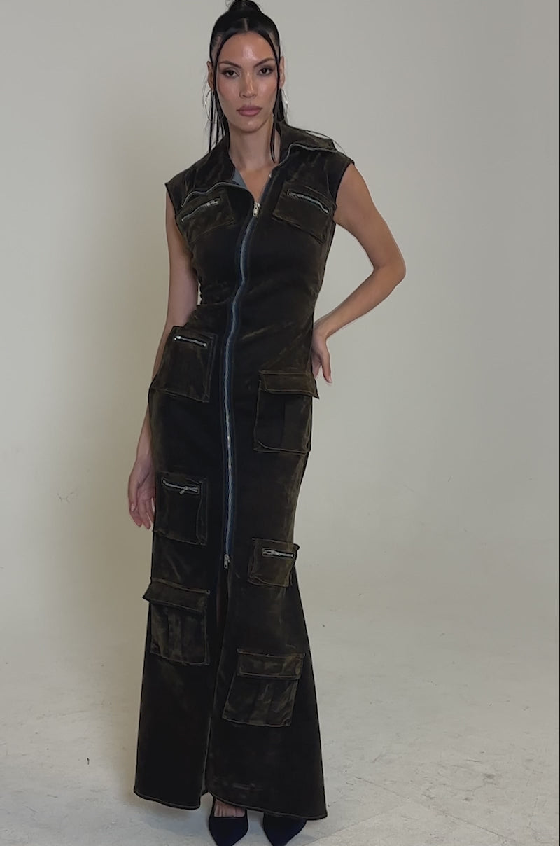 A video of a woman modeling the NOW YOU SEE ME VELVET DENIM MAXI DRESS, a long, sleeveless dress with a front zipper and cargo pockets. She wears dark heels and has long hair in two small top knots against a plain, light background.
