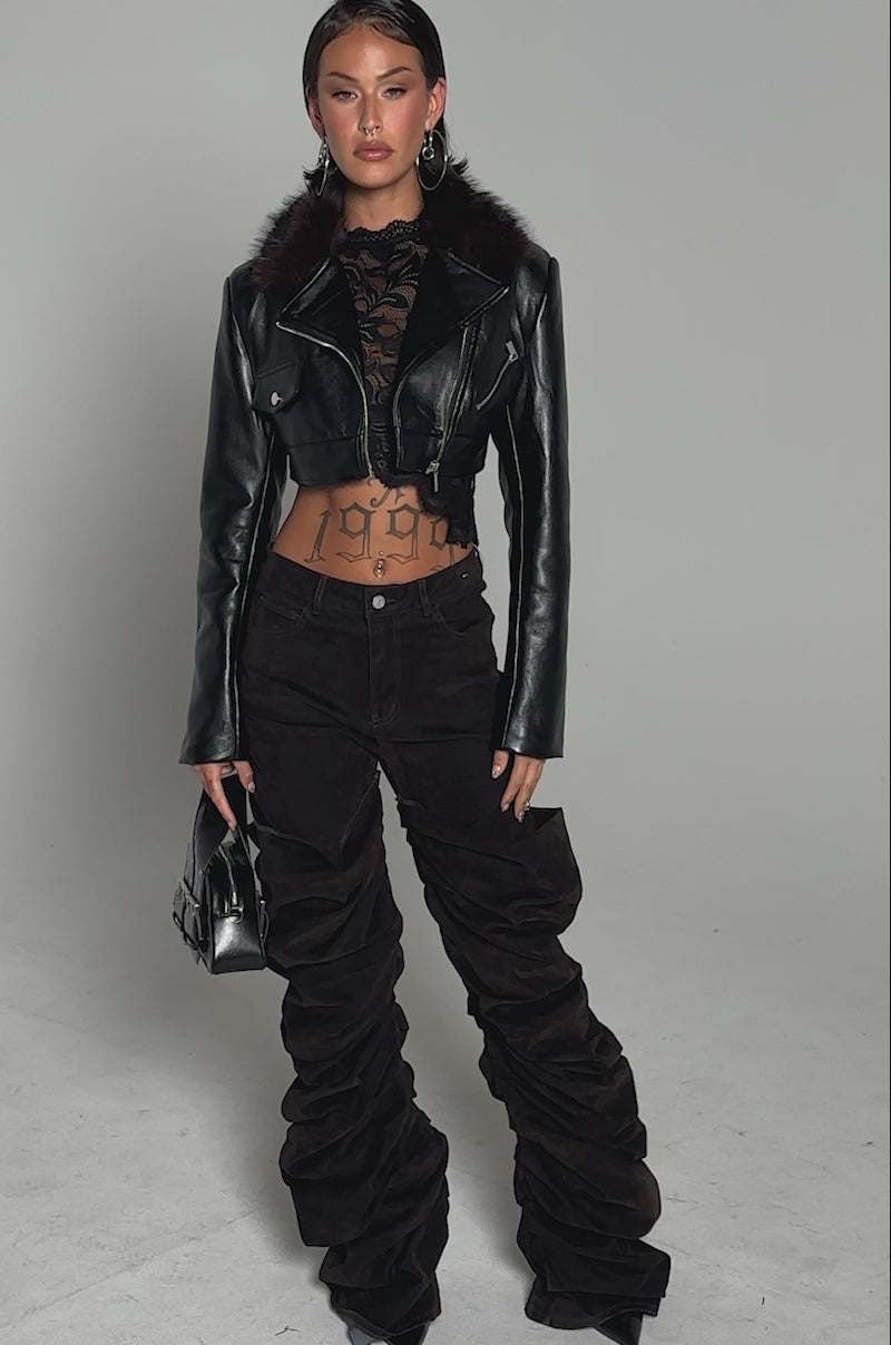 A video of a person modelling the VICTORIA FUR TRIM MOTO jacket with fur trim, styled with a sheer black lace top, brown ruched pants, pointed black shoes, and a small black handbag against a plain background.