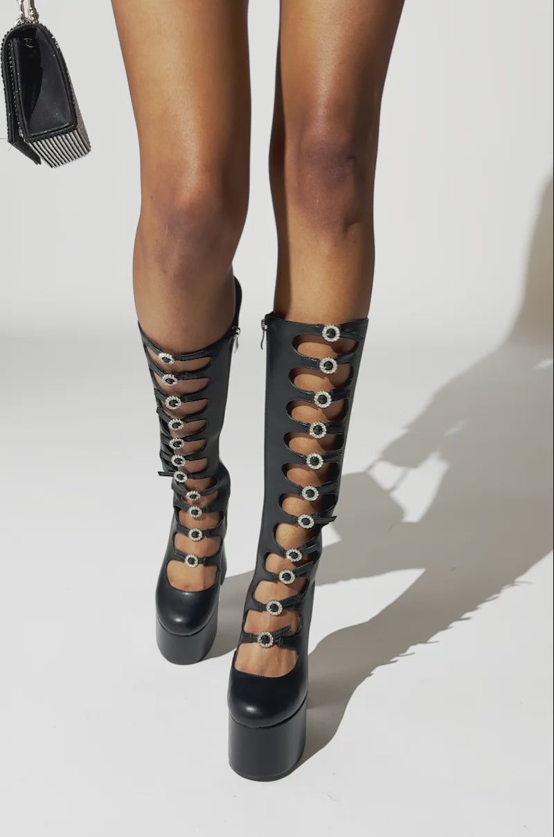 A video of a person wearing the AZALEA WANG TOMLIN BLACK BOOT—knee-high, black boots with platform soles, multiple cut-outs, and silver buckle details—standing on a light background.