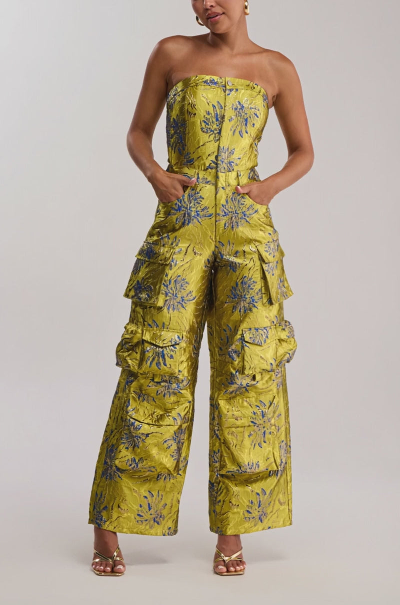 Video of woman modeling the NIKKI BROCADE JUMPSUIT IN GREEN, a strapless floral wide-leg jumpsuit with cargo pockets, styled with gold heels and hoop earrings.