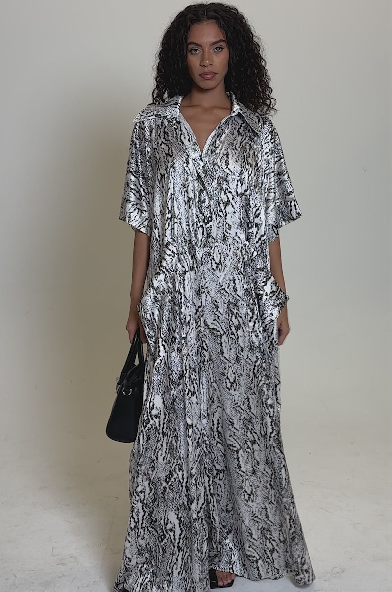Video of woman modeling the SHE'S RICH Oversized Satin Jumpsuit in Snake Print, featuring short sleeves and a collar. She holds a black handbag and stands against a plain, light background.