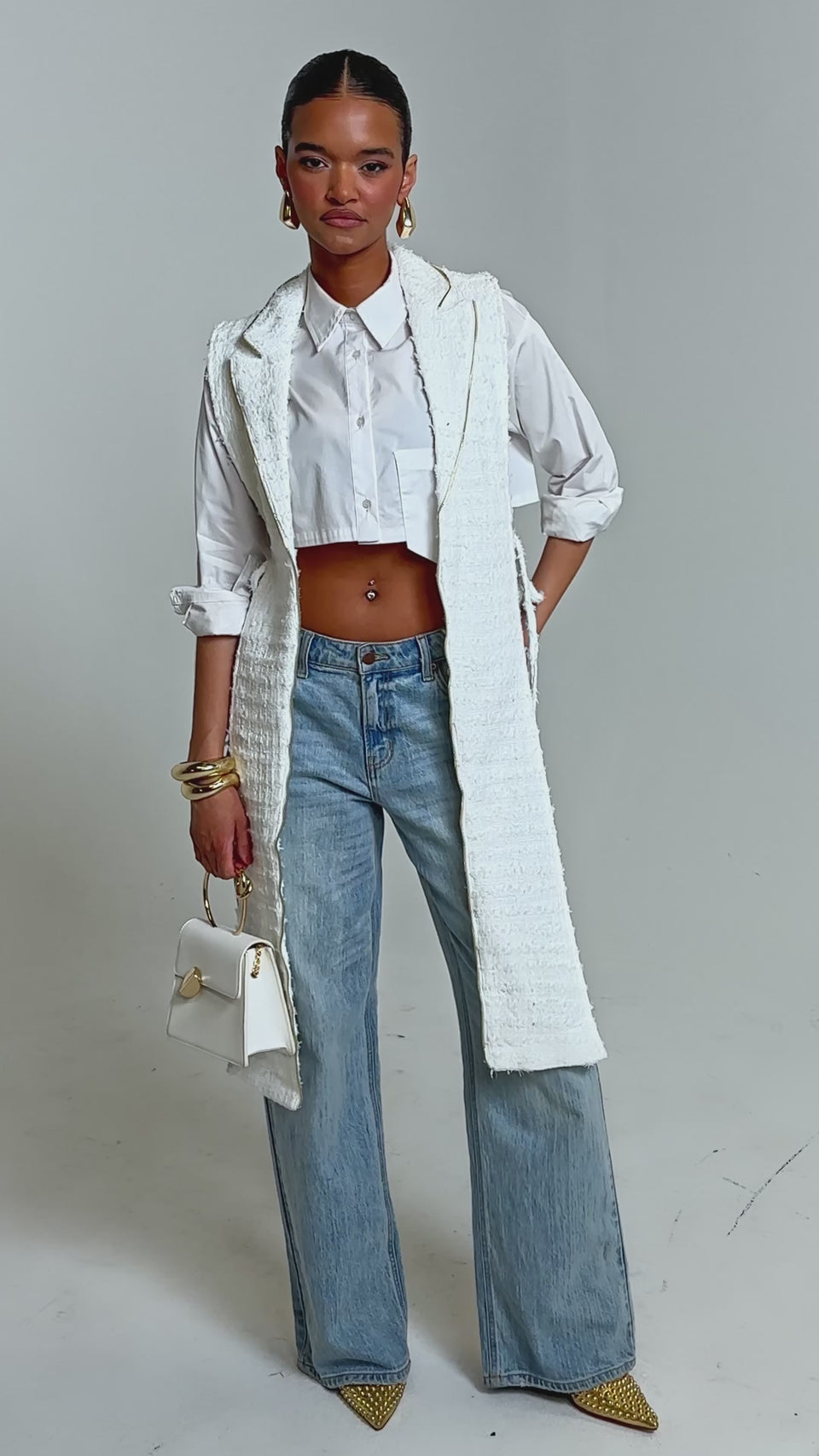 Video of woman modeling the HONEY HONEY SPLIT VEST IN TWEED. She accessorizes with gold jewelry, carries a small white handbag, and stands against a neutral background.