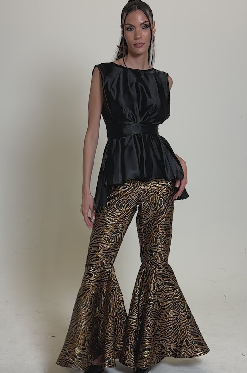 Video of woman modeling the GET IN MOTION BROCADE FLARE TROUSER, paired with a sleeveless black peplum top and black heels. She has long, straight hair and stands against a plain gray background.