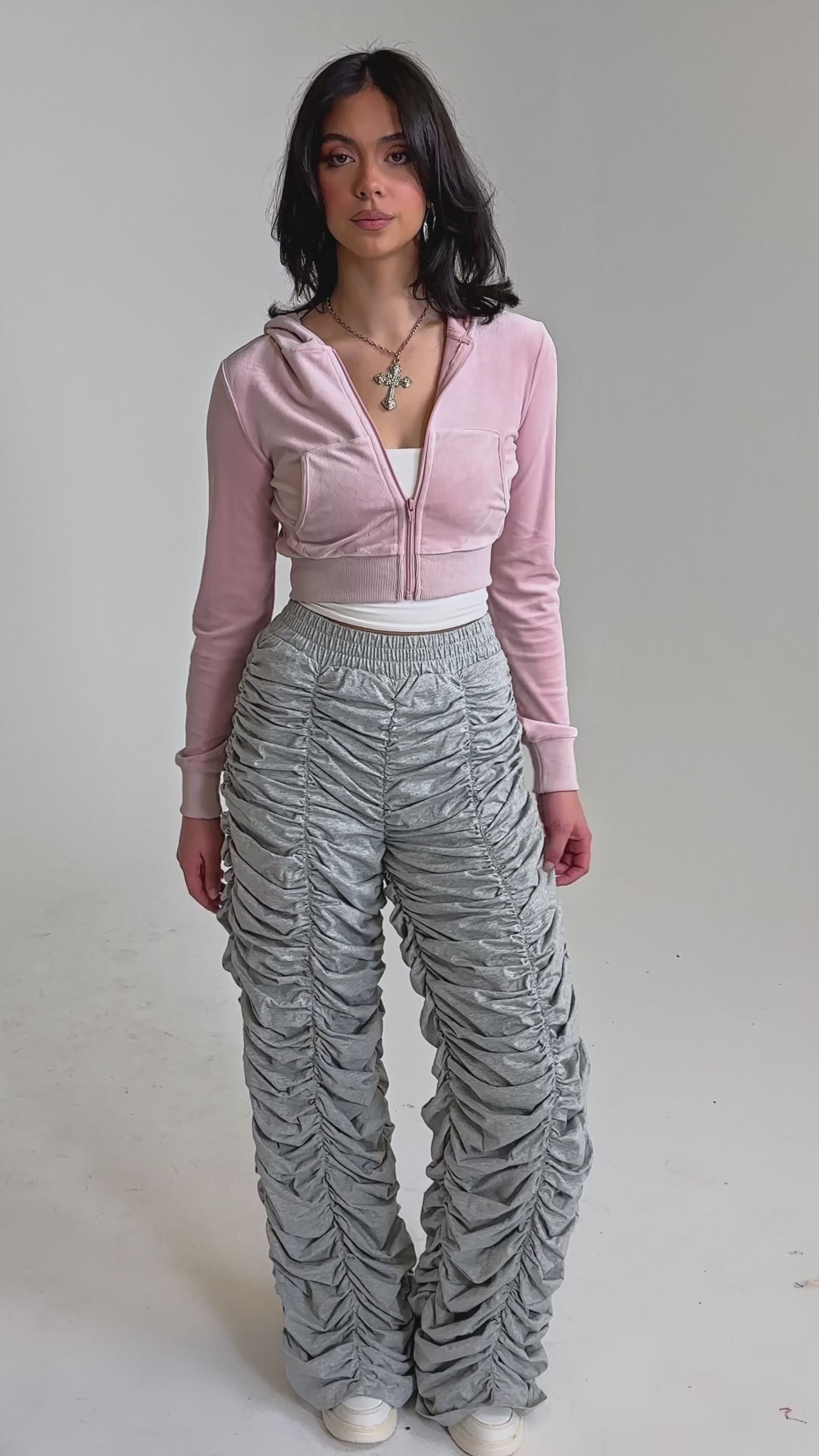 A video of a woman wearing an AKIRA Label light pink cropped zip-up hoodie, IM THAT TYPE RUCHED JOGGER heather grey joggers, white sneakers, and layered necklaces.