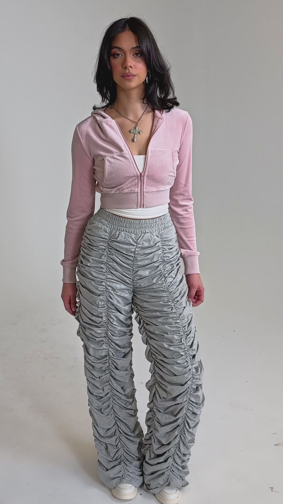 A video of a woman wearing an AKIRA Label light pink cropped zip-up hoodie, IM THAT TYPE RUCHED JOGGER heather grey joggers, white sneakers, and layered necklaces.