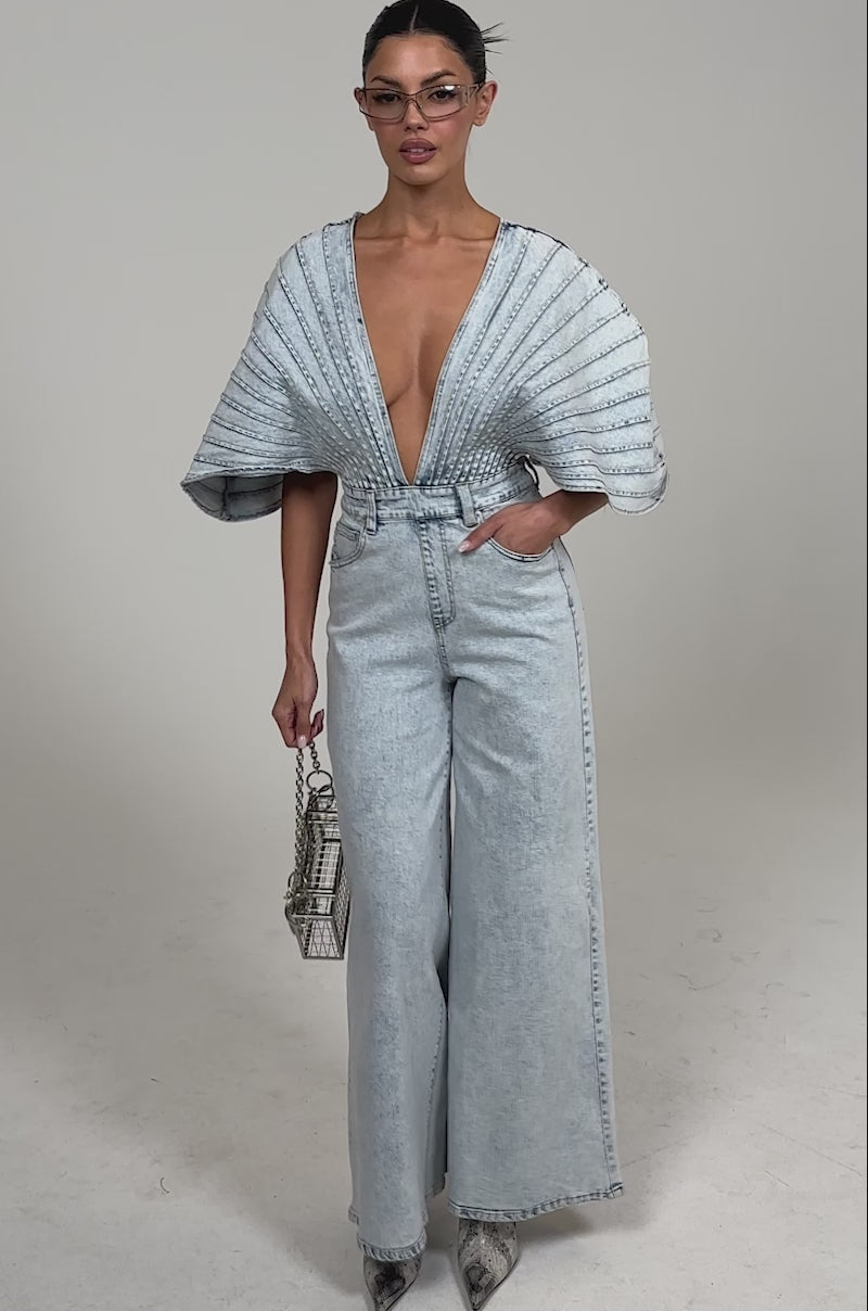 Video of woman modeling the BORN TO STAND OUT DENIM JUMPSUIT with pleated bodice and dramatic sleeves, gray snakeskin boots, geometric glasses and a metallic cube-shaped handbag.