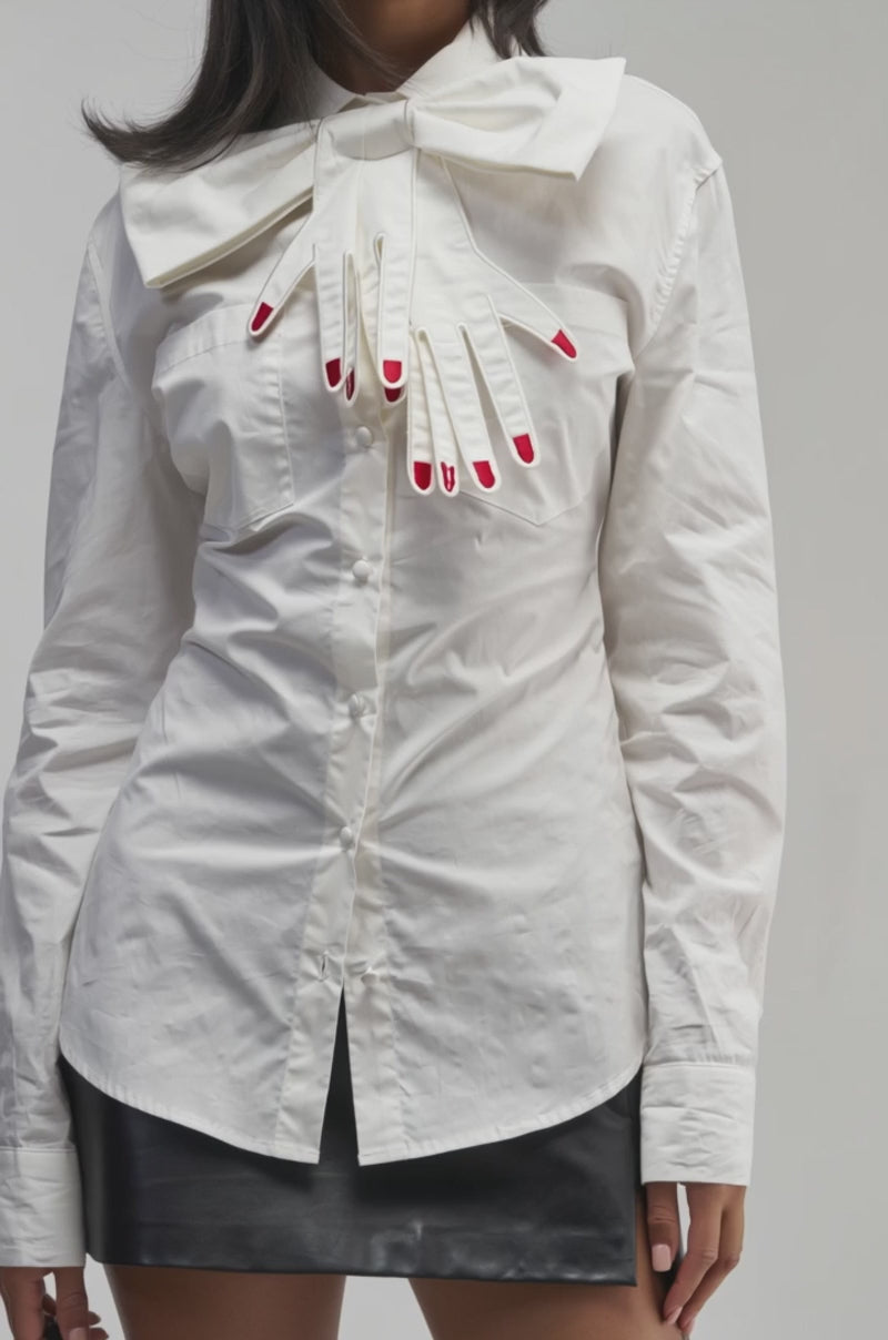 Video of a woman modeling the HANDS OFF POPLIN BLOUSE, styled with a large hand-shaped bow tie featuring red nails. 