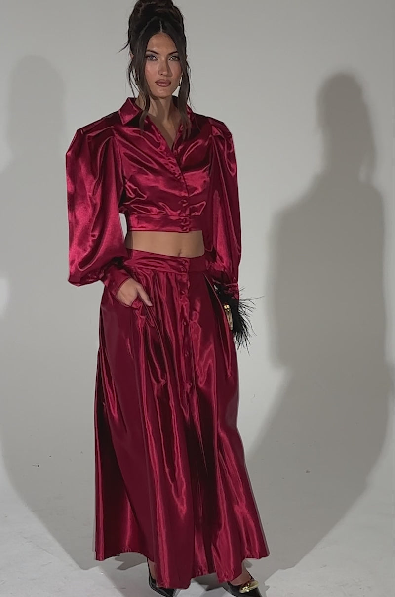 A video of a woman posing against a plain background in the CANDICE OPEN BACK SATIN BUTTON DOWN SHIRT, styled in deep red with long sleeves, paired with a matching skirt, black pointed heels, and a small feathered clutch.