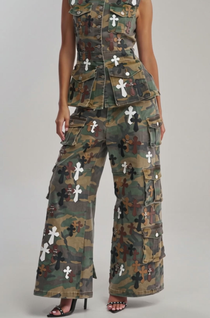 Video of woman modeling the DOWN AND DIRTY CAMO BUTTON DOWN, a sleeveless camouflage top with brown, black, and white cross patches, standing against a plain background, exuding bold streetwear style.