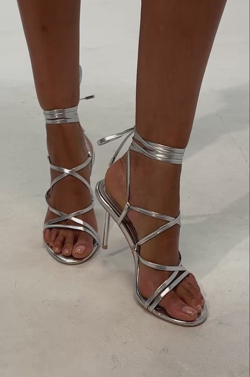 Video of woman modeling the AZALEA WANG KATRIONA Silver Patent Metal Heel Lace Up Sandals. These sandals feature slim, metallic stiletto heels and thin silver straps that lace up around the ankles, making them an ideal choice for formal occasions. 