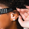 Close-up of a person wearing pearl-studded black sunglasses and a SEXY STUD EARRING SILVER. The person's ear and part of their cheek are visible, showing areas of vitiligo. A hand with manicured nails gently touches their curly hair.