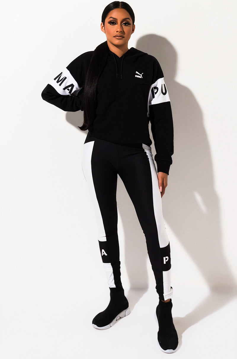 A woman wearing a black and white Puma tracksuit stands against a white background. Her outfit includes a black hoodie with white text on the sleeves and SEXY SKINNY LEGGING, which are black with white panels along the sides and the letter "P" near the ankles. She wears black sneakers.