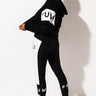 A person wearing a black and white athletic outfit is standing in a side profile pose. The outfit includes a black hoodie with "PUMA" written on the sleeve, SEXY SKINNY LEGGING featuring the Puma logo, and black athletic shoes. The background is plain white.