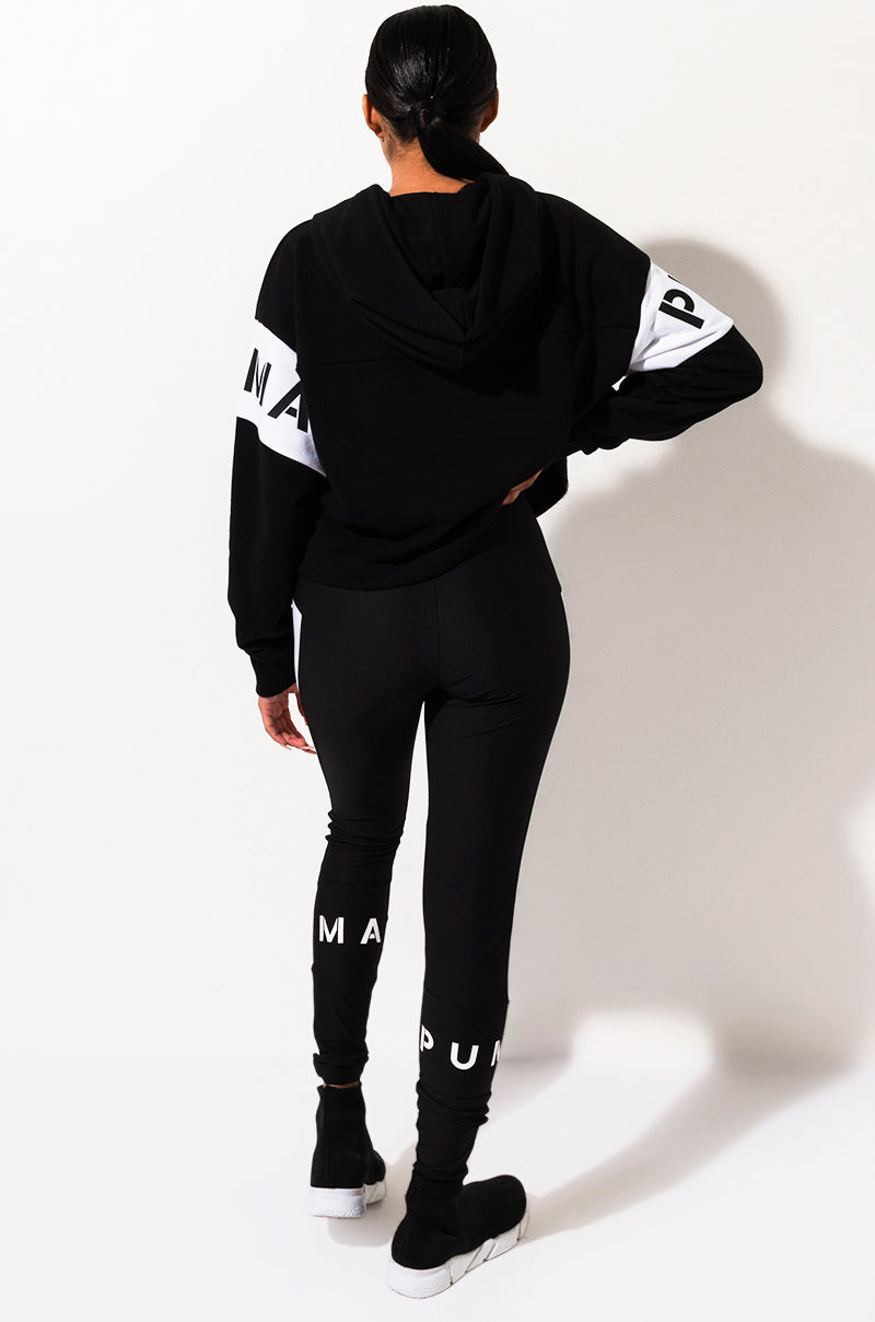 The image shows a person facing away from the camera, wearing a black hooded jacket with white text on the sleeves, SEXY SKINNY LEGGING in black with white text on the lower legs, and black sneakers with white soles. The person is standing against a plain white background.