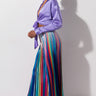 A woman stands against a plain background wearing a shiny lavender tied-front blouse and the SEXY JOGGER PANT with colorful rainbow stripes. She has large hoop earrings and clear high-heeled sandals, and her hair is neatly tied back.