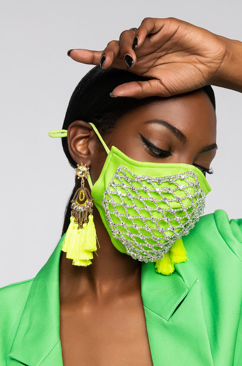 A person is wearing the READY WHEN YOU ARE RHINESTONE FASHION FACE COVER, accentuated with a beaded net pattern. They have also accessorized with neon green tassel earrings. With their hand partially raised to display black nail polish, they complete the look with a vibrant green outfit, achieving a bold and fashionable appearance.