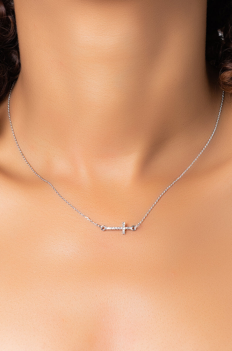 Close-up of a person wearing the NECKLACE, a delicate silver chain featuring a small horizontal cross pendant. The blurred background accentuates the contrast against the person's skin, highlighting the elegant necklace.